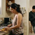 mãe cozinhando na cozinha enquanto filho adolescente observa da porta com celular na mão