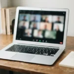 Notebook aberto em videochamada sobre a mesa de trabalho em casa, com bloco de notas e caneta ao lado, representando como ser mais produtiva em reuniões de trabalho remoto